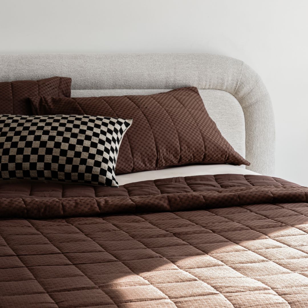 A modern bed with a light-gray upholstered headboard, brown quilted bedding, brown pillows, and a black-and-white checkered accent pillow against a plain white wall.