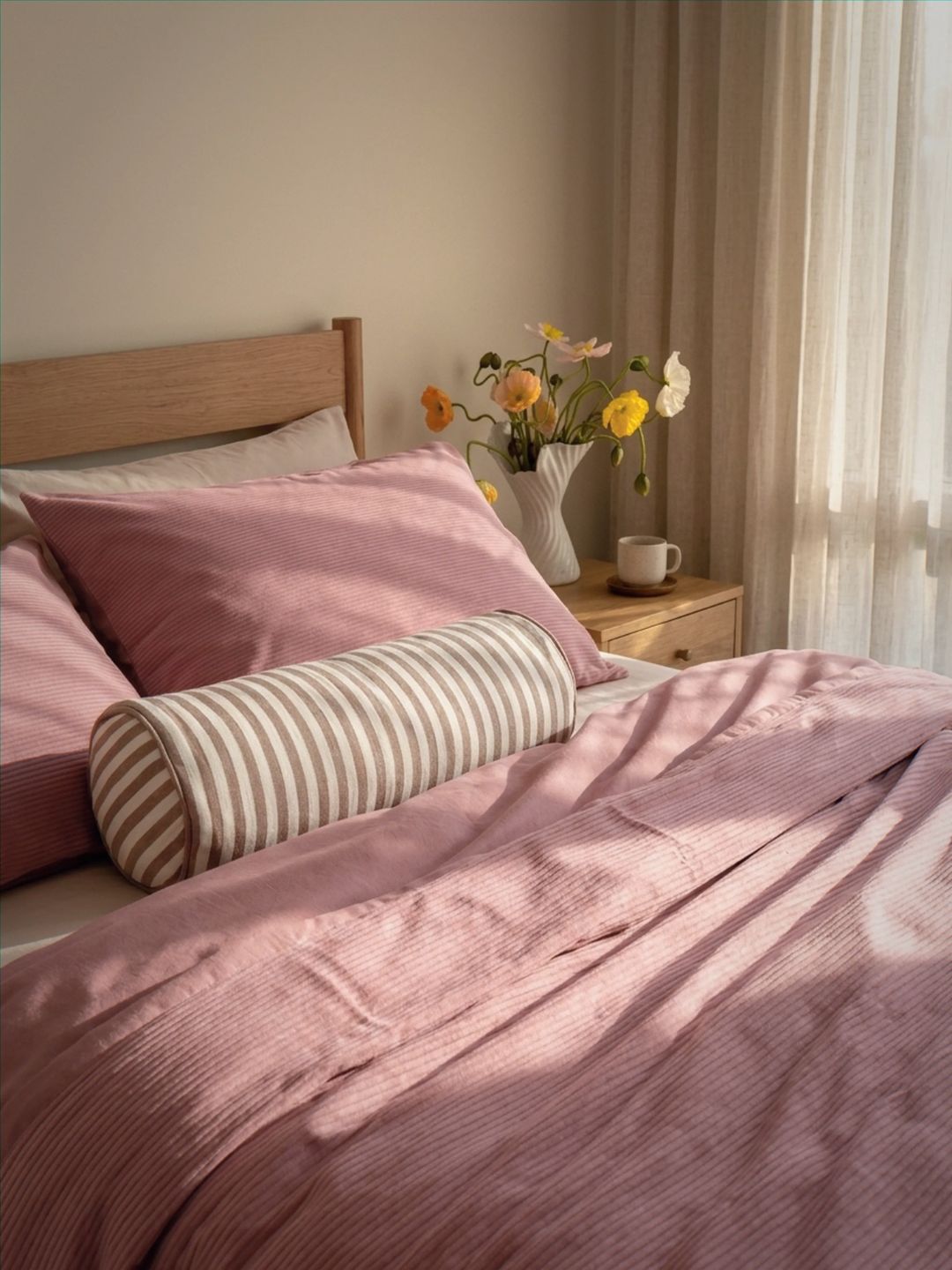 Bedroom with pink bedding, striped bolster pillow, and a vase of flowers on a nightstand.
