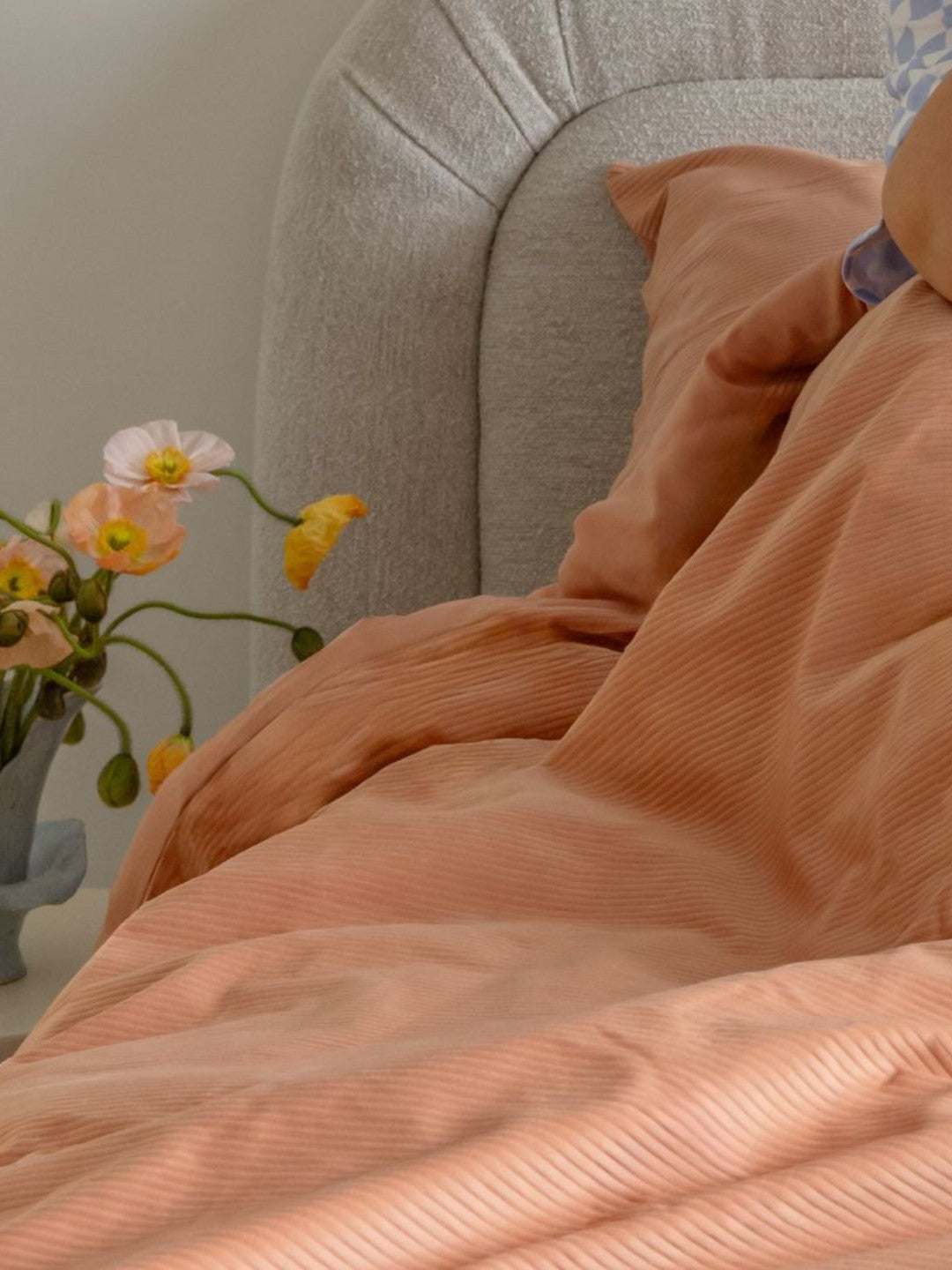 Orange comforter on a bed with a vase of flowers on a table in the background.