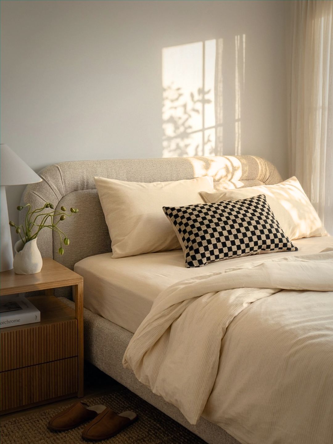 A neatly made bed in a softly lit bedroom, dressed in cream bedding with a black-and-white checkered accent pillow. A wooden bedside table with a white lamp and vase, a pair of brown slippers, and a window shadow on the wall are visible.