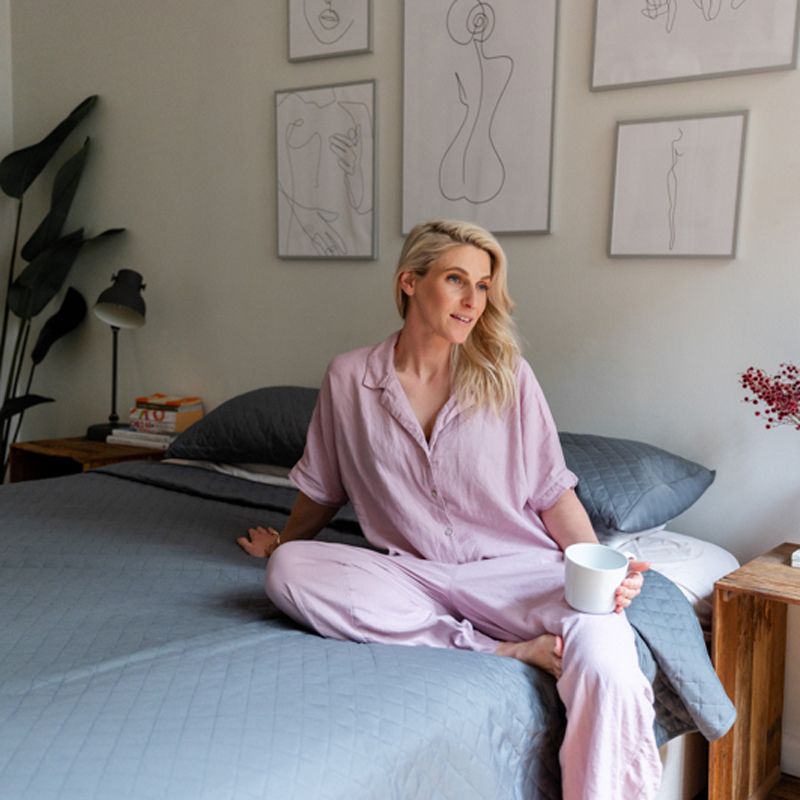 A neatly made bed features the Morgan and Reid Argyle Shade Coverlet Set in dark gray with matching pillows, creating a contemporary vibe. Above, three abstract art pieces hang, while a wooden table with red flowers sits to the right.