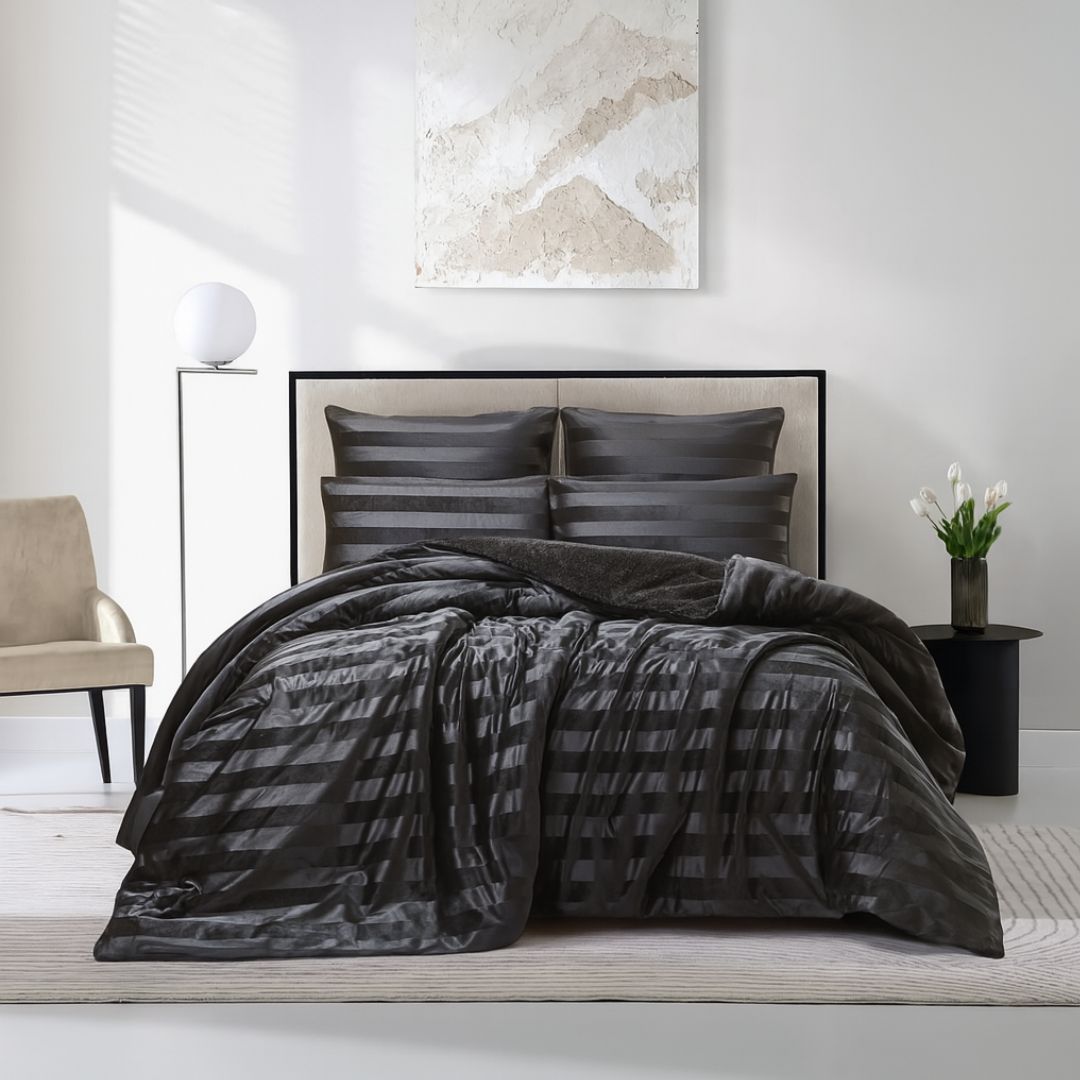 A modern bedroom features the Morgan and Reid Black Velvet Stripe Snuggle Fleece Comforter Set on a neatly made bed with striped pillows, neutral walls, abstract art, a beige chair, round black side table with white flowers, and a lamp.