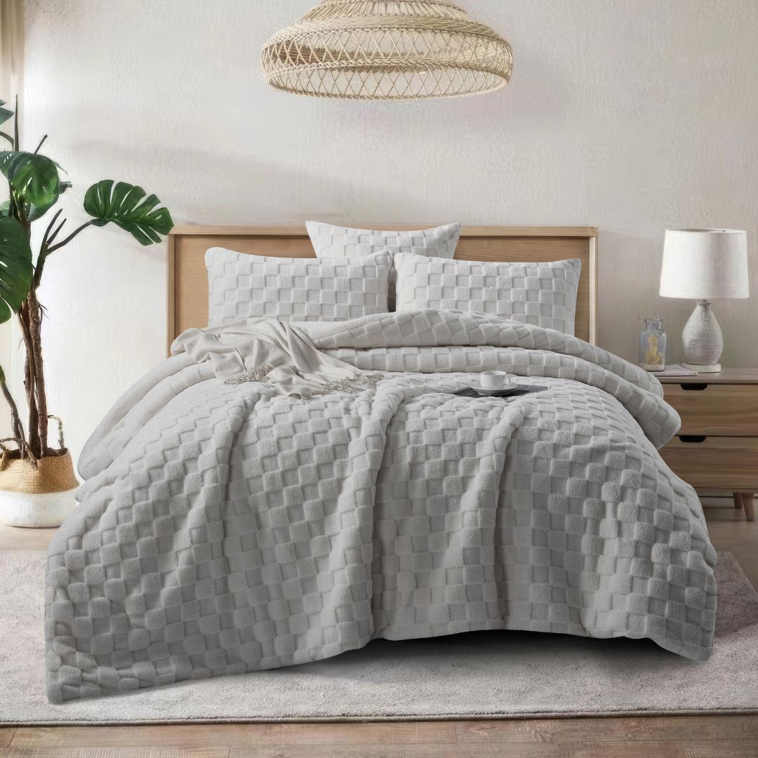 A neatly made bed features the Morgan and Reid Checkered Silver Snuggle Fleece Comforter Set, with matching pillows, a wooden headboard, nightstand with lamp and bottles, a large plant, and a woven pendant light for lasting luxury.