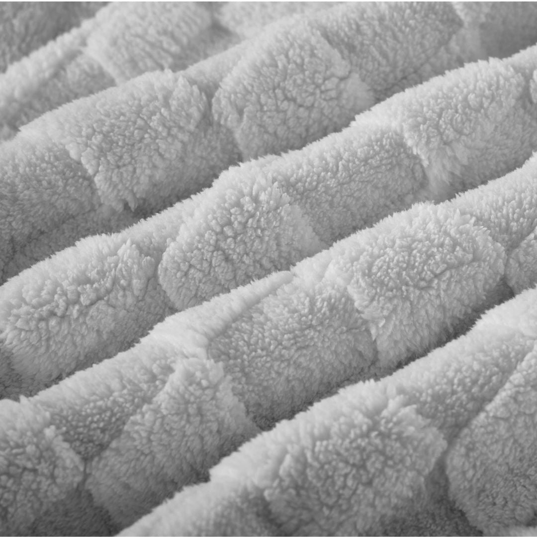 A neatly made bed features the Morgan and Reid Checkered Silver Snuggle Fleece Comforter Set, with matching pillows, a wooden headboard, nightstand with lamp and bottles, a large plant, and a woven pendant light for lasting luxury.