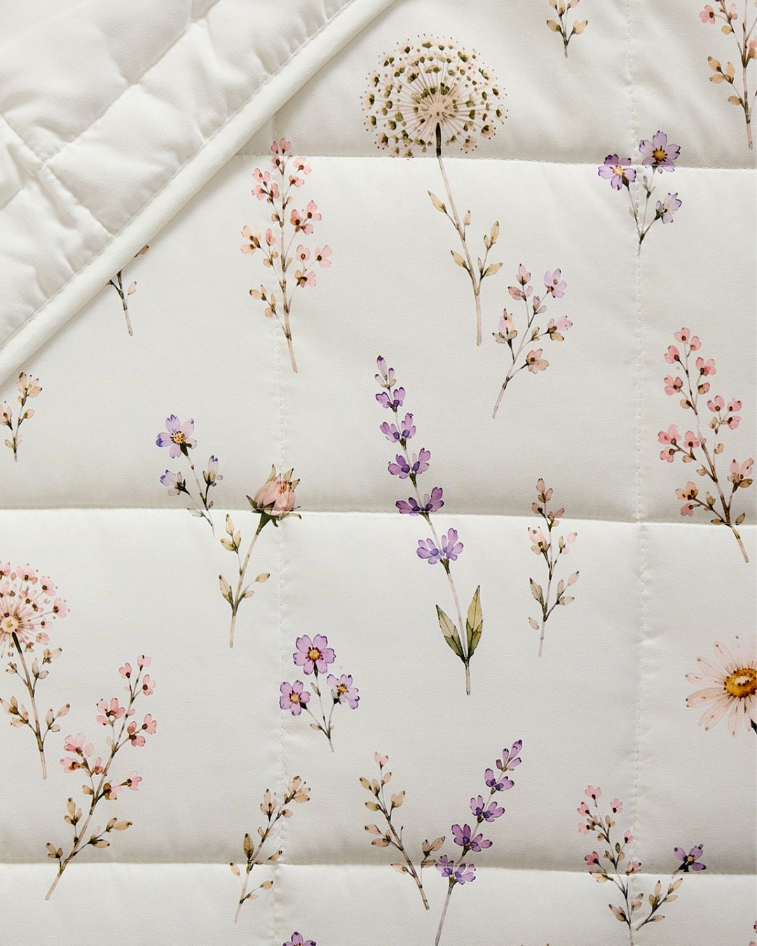 A close-up of a quilted white or off-white bedding surface with a scattered floral print in soft pink, purple, and muted green tones.