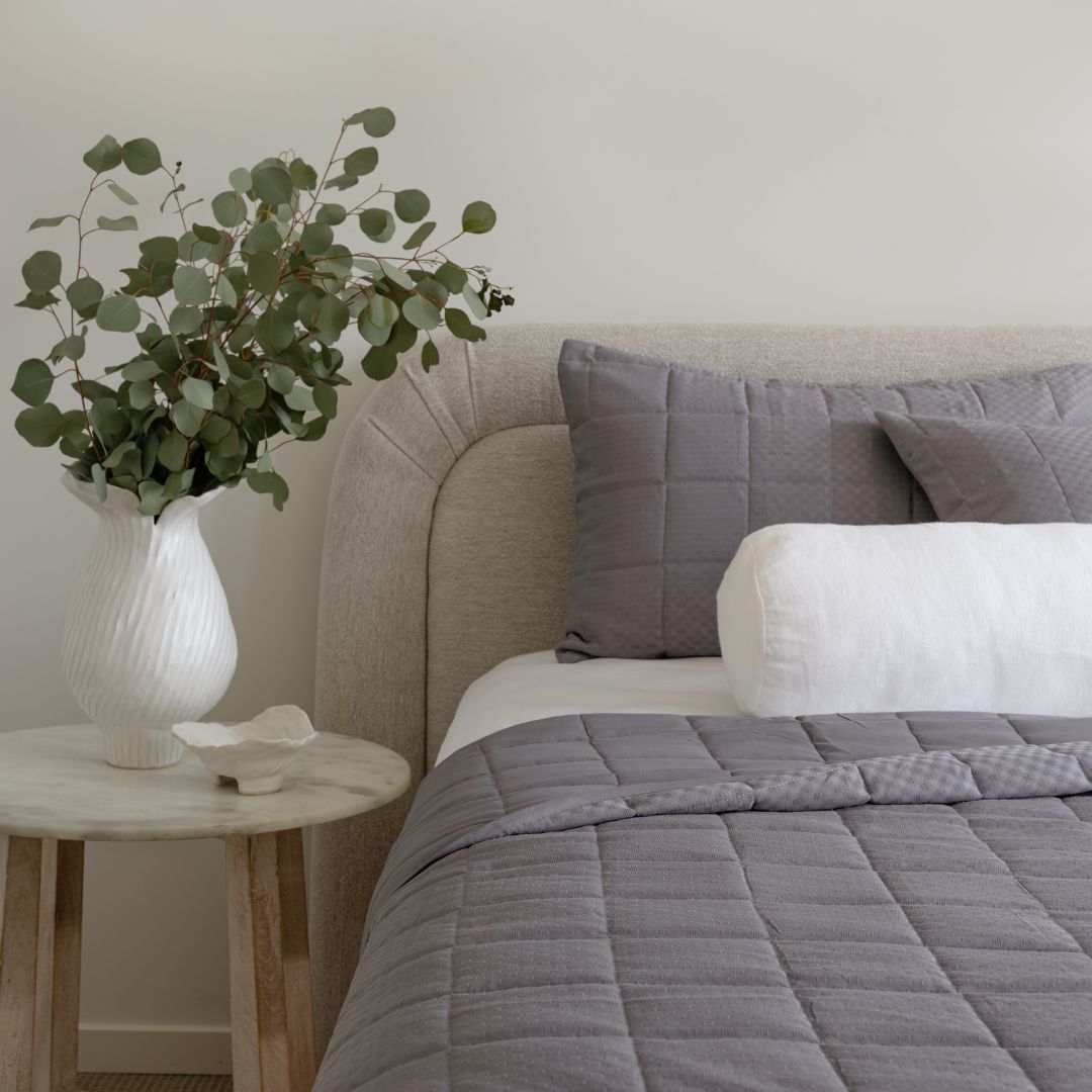 Bedroom with a bed featuring gray bedding, a vase with greenery on a nightstand, and a neutral wall.