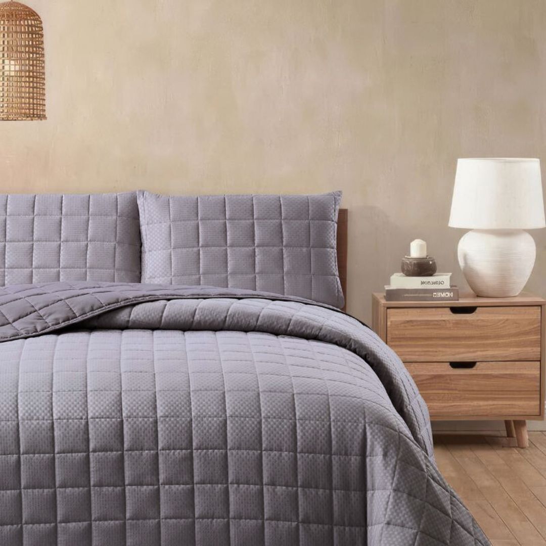 Gray quilted bedding set on a bed with a wooden nightstand and lamp in a bedroom setting.