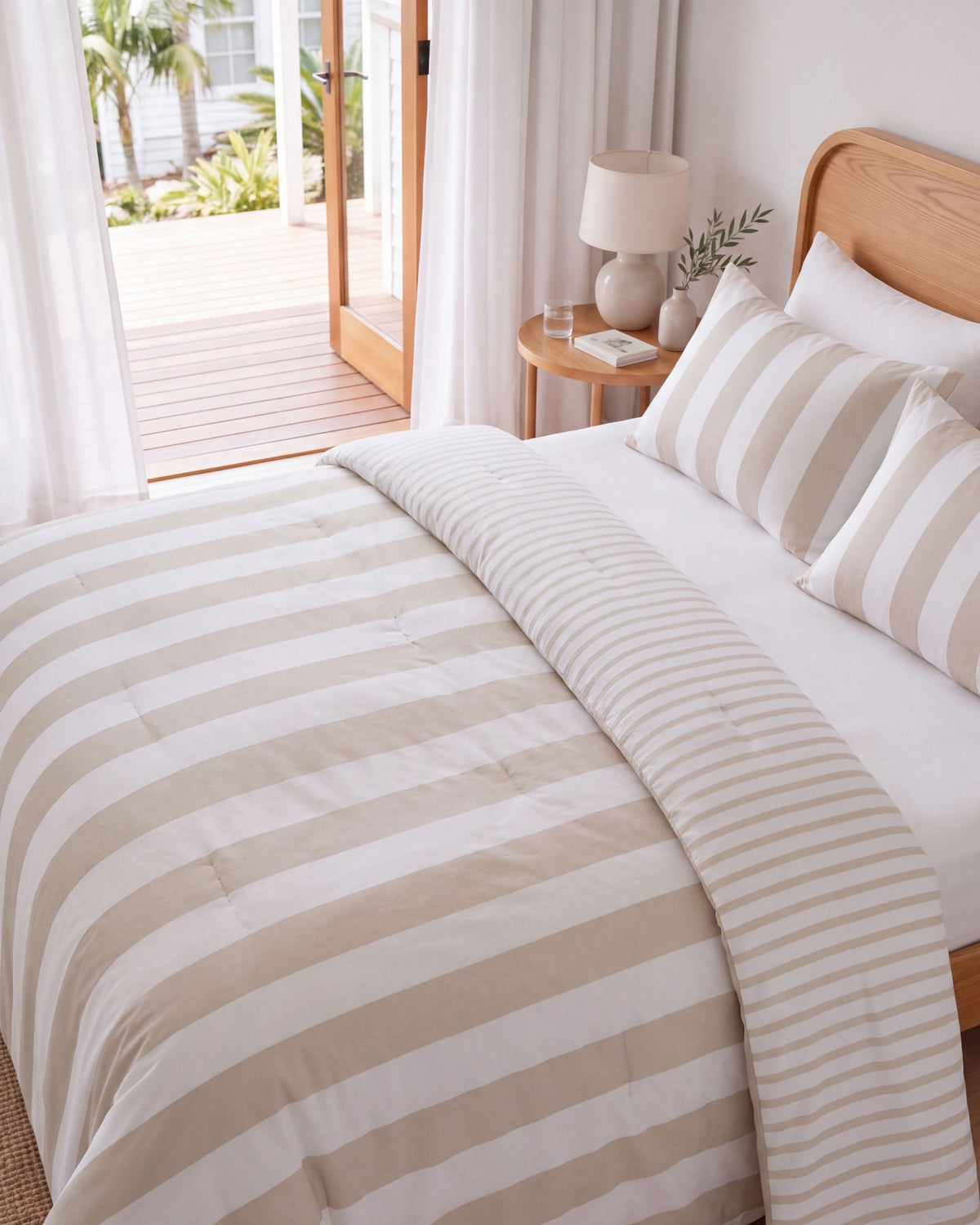 A neatly made bed in a bright bedroom with beige and white striped bedding, including a duvet cover and matching pillow shams, a wooden headboard, a bedside table with a lamp, and an open door leading to a wooden deck.
