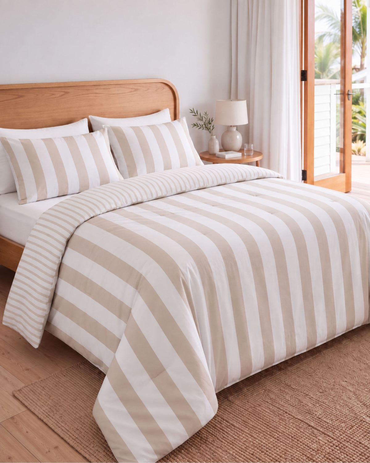 A neatly made bed in a bright bedroom setting, featuring a beige and white striped duvet cover and matching pillow shams. The bed has a light wood headboard, a small wooden nightstand with a lamp, and a window with curtains and glass doors in the background.