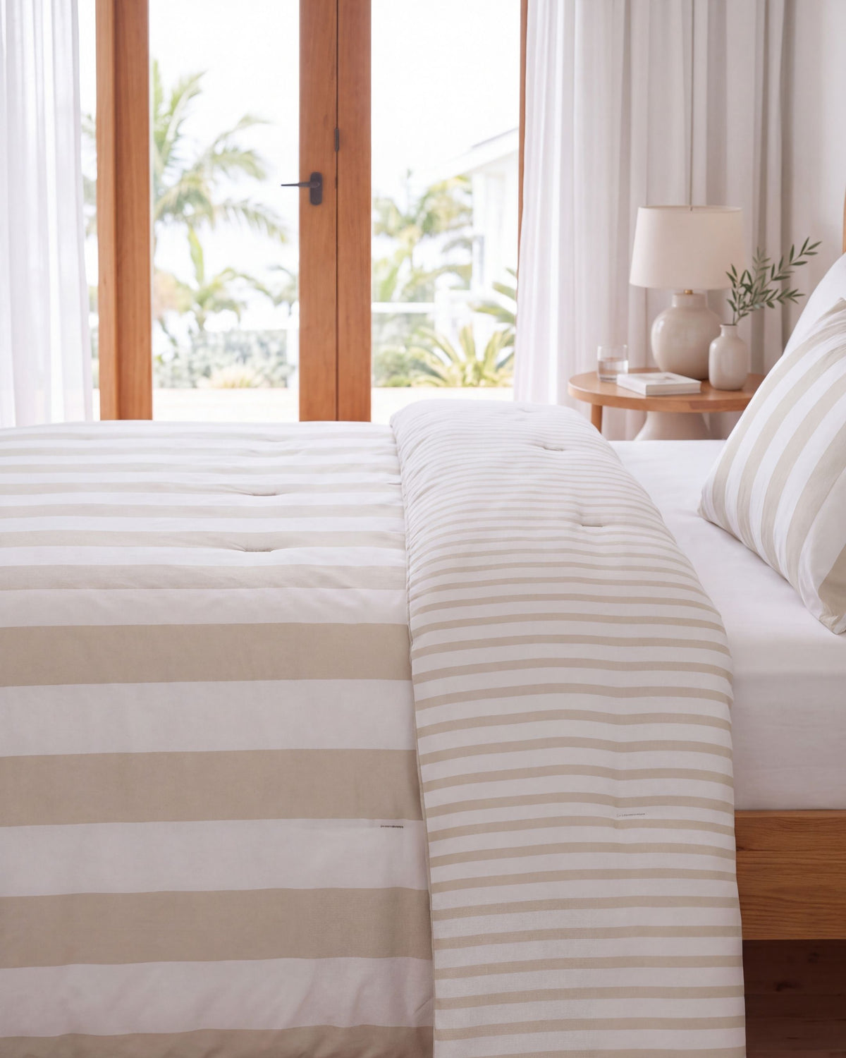 A neatly made bed in a bright bedroom with white and beige striped bedding, including a duvet cover and matching pillow shams, set against a background of wooden doors, sheer curtains, and a bedside lamp.