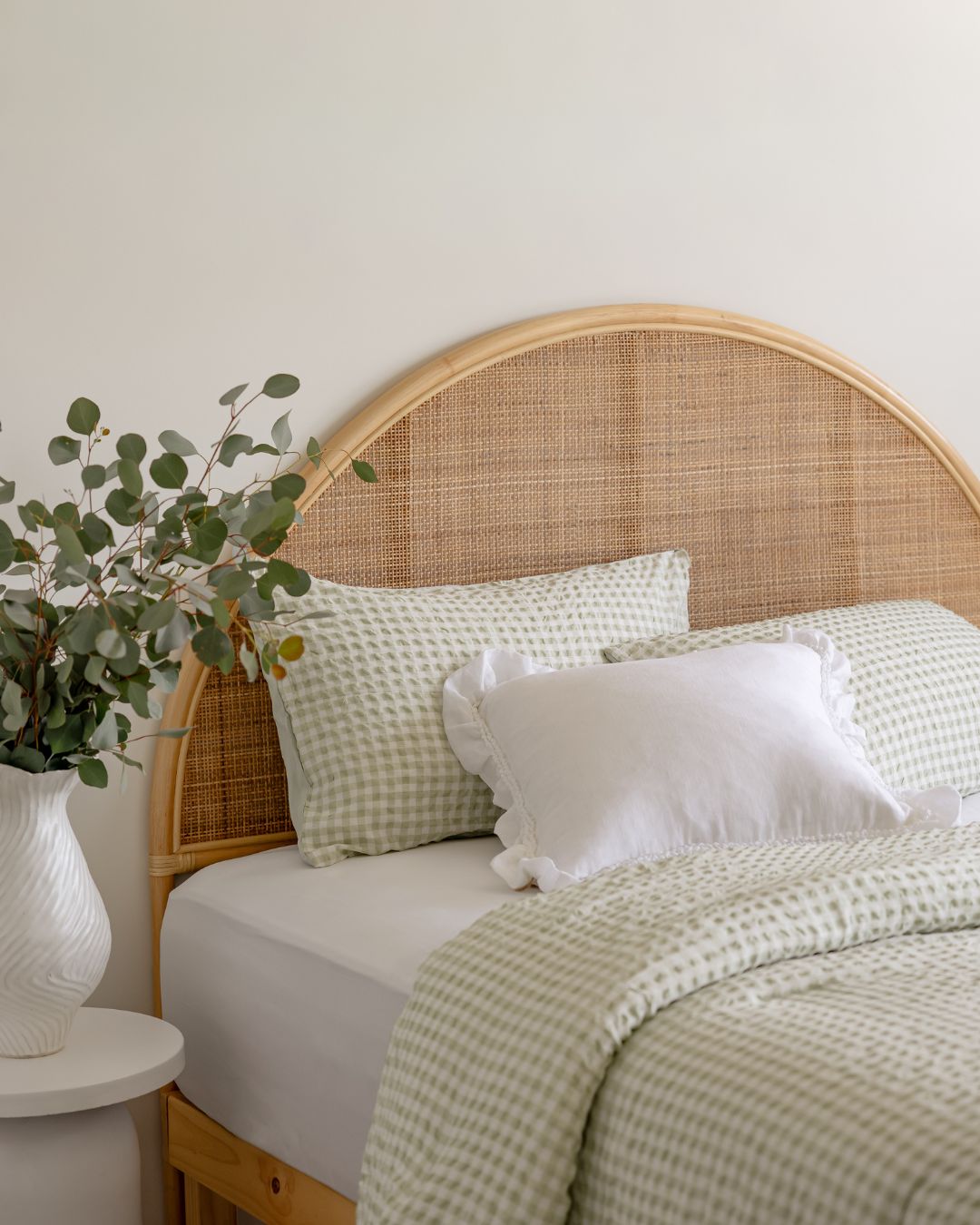 Bed with wicker headboard and green bedding, next to a plant on a white side table.