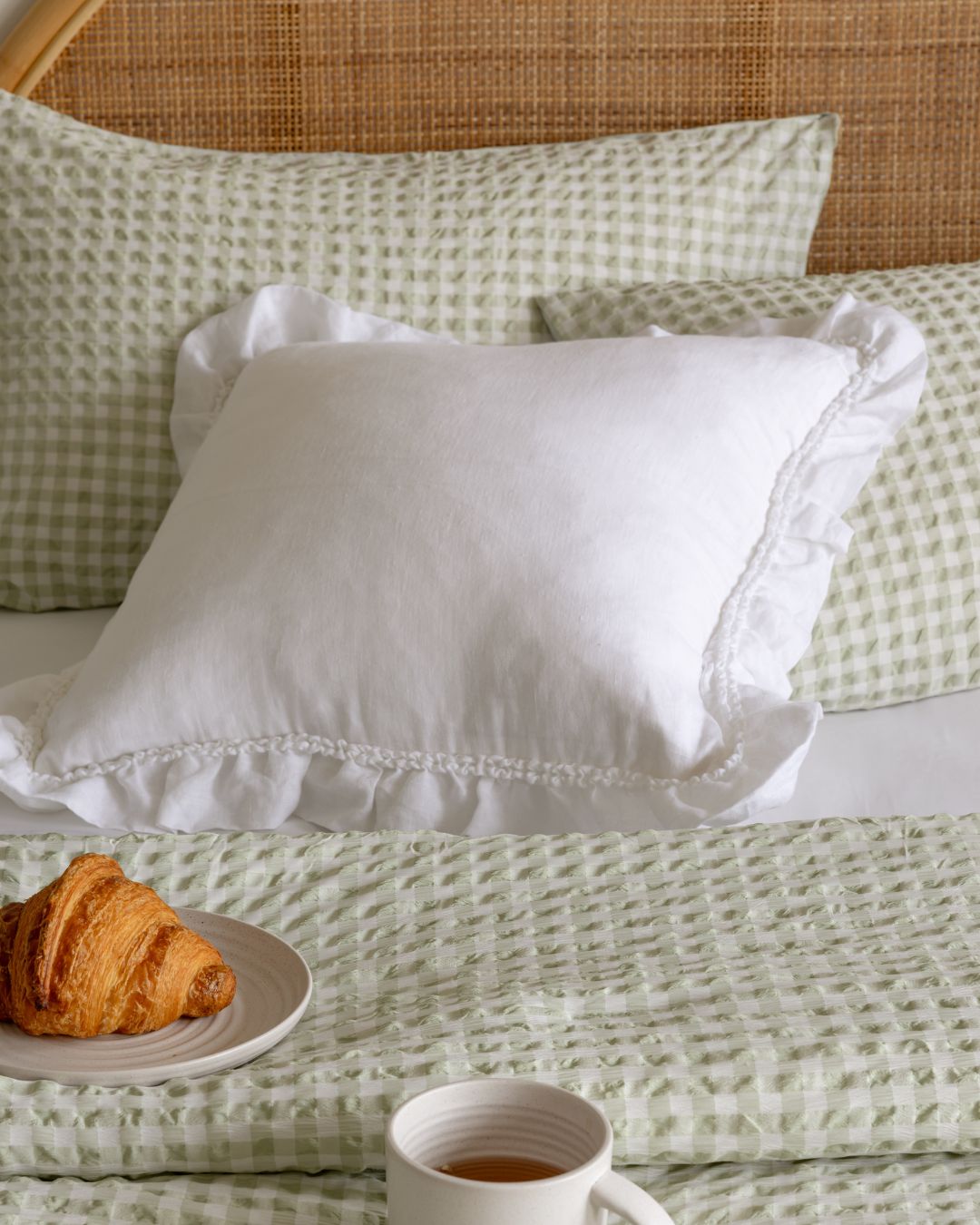 White pillow on a bed with green checkered sheets and a croissant on a plate.