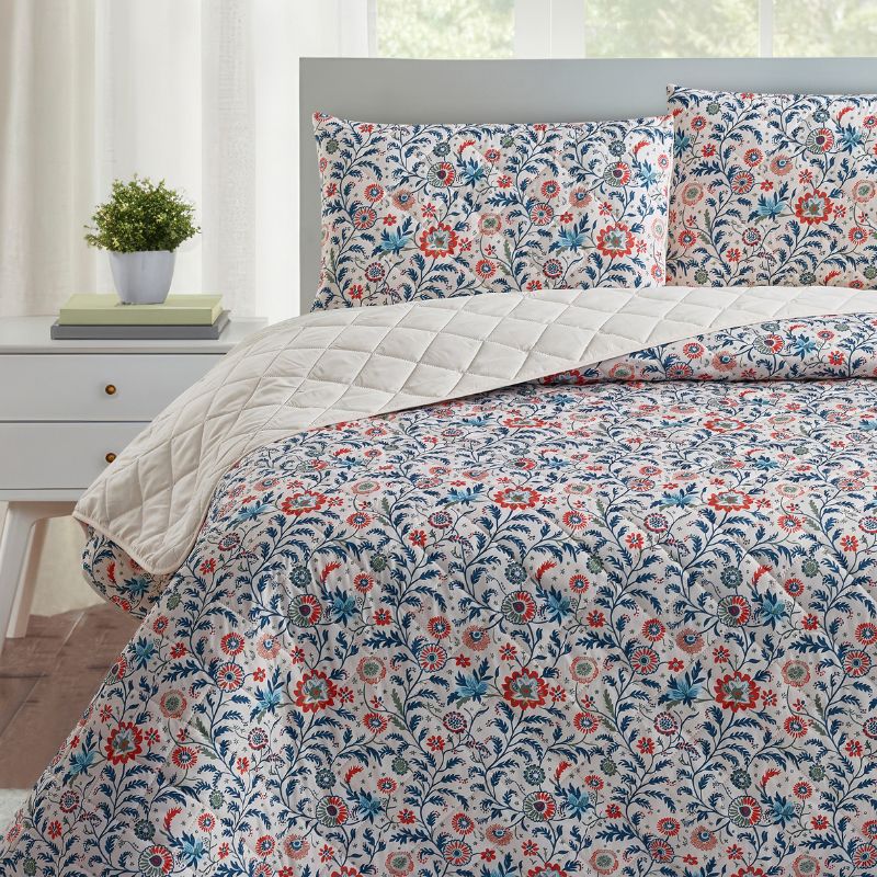 A bright bedroom features the Morgan and Reid Indian Floral Coverlet Set with matching pillow shams, complemented by white walls, large windows, a small lamp, plant, and alarm clock on bedside tables.