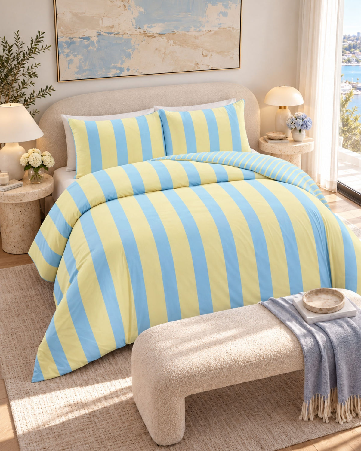 A bed dressed in a yellow and light blue vertically striped duvet cover and matching pillow shams, styled in a bright, modern bedroom with bedside tables, lamps, a bench, and a window view.