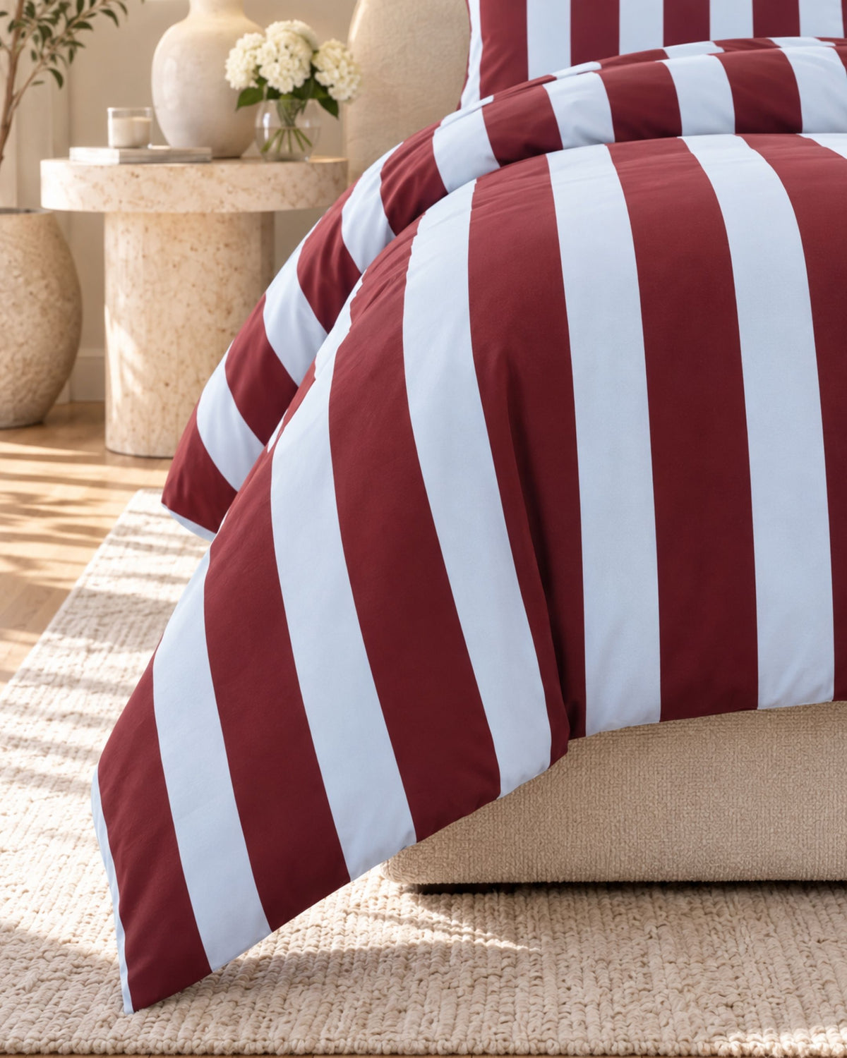 A close-up of a bed dressed in a burgundy and white vertically striped duvet cover and pillowcase, styled in a warm, sunlit bedroom with a beige rug and side table.