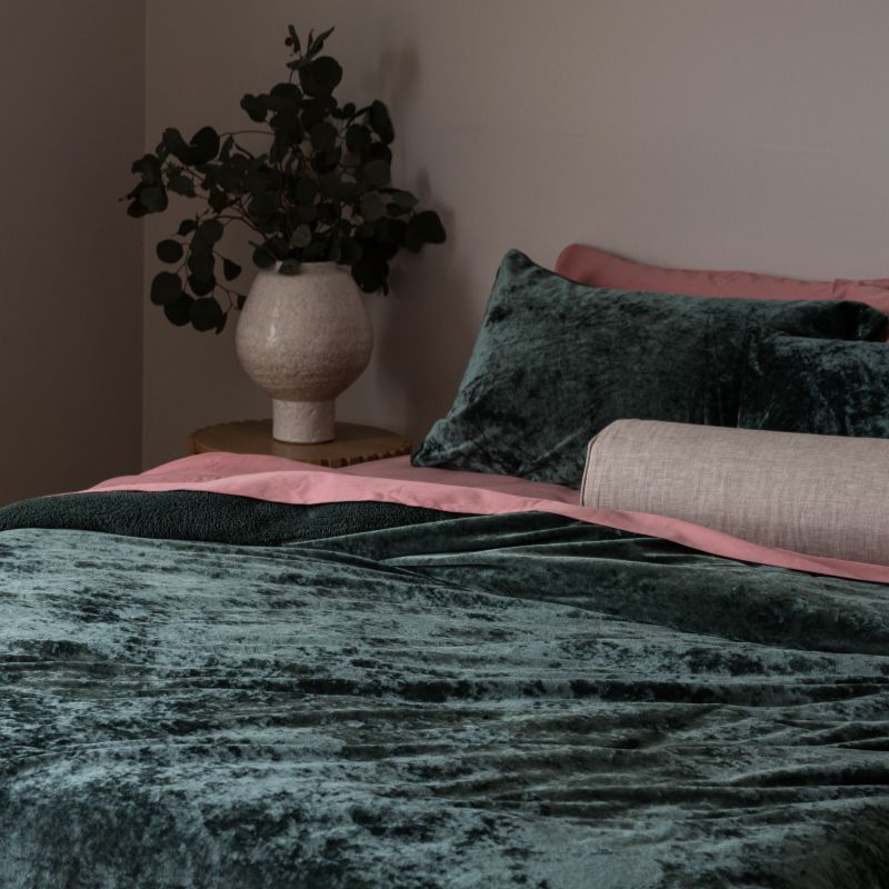 Bed with green velvet bedding and pink pillows, vase with plants on nightstand.