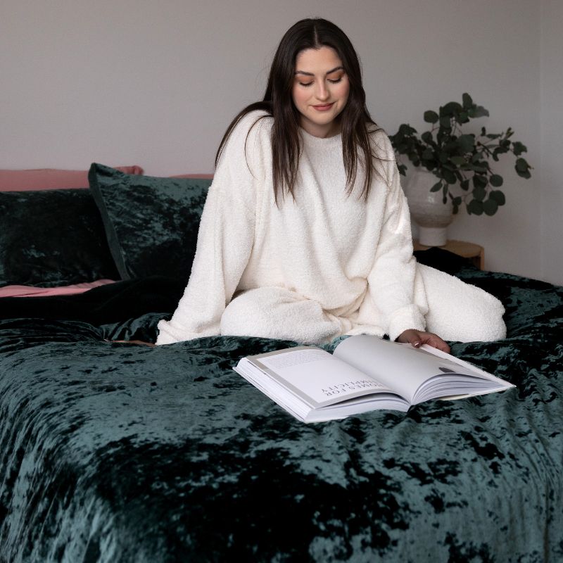 Woman in a white robe reading a book on a dark green bedspread.