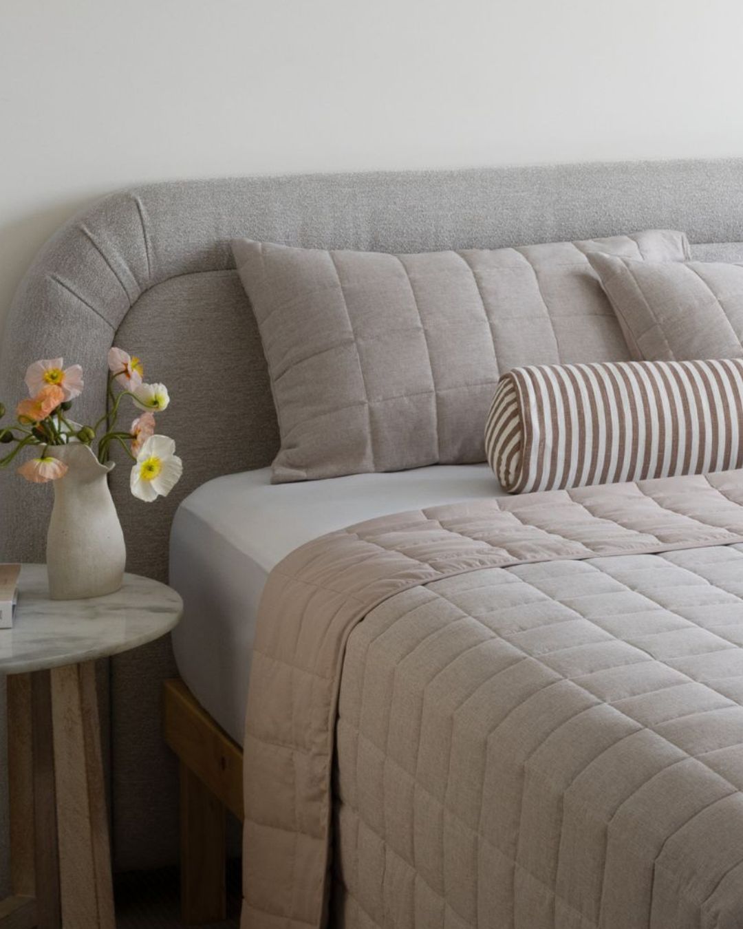 Neatly made bed with beige bedding and striped pillow, side table with flowers in a room.