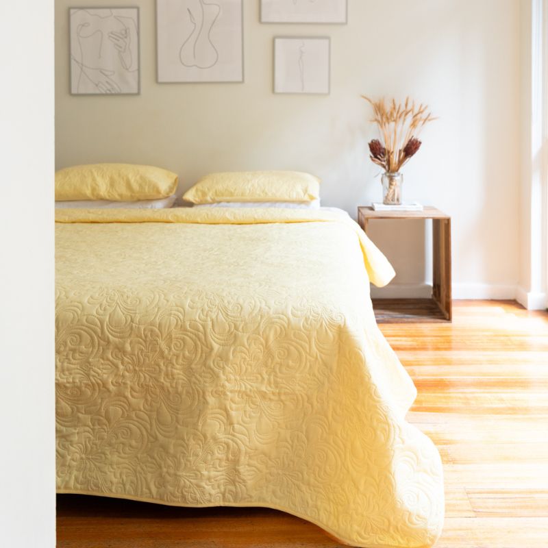 The Morgan and Reid Serene Butter Coverlet Set by Morgan and Reid, featuring a yellow coverlet and two matching pillows, brightens the room. A wooden nightstand with dried flowers and minimalist line art complete the cozy look.