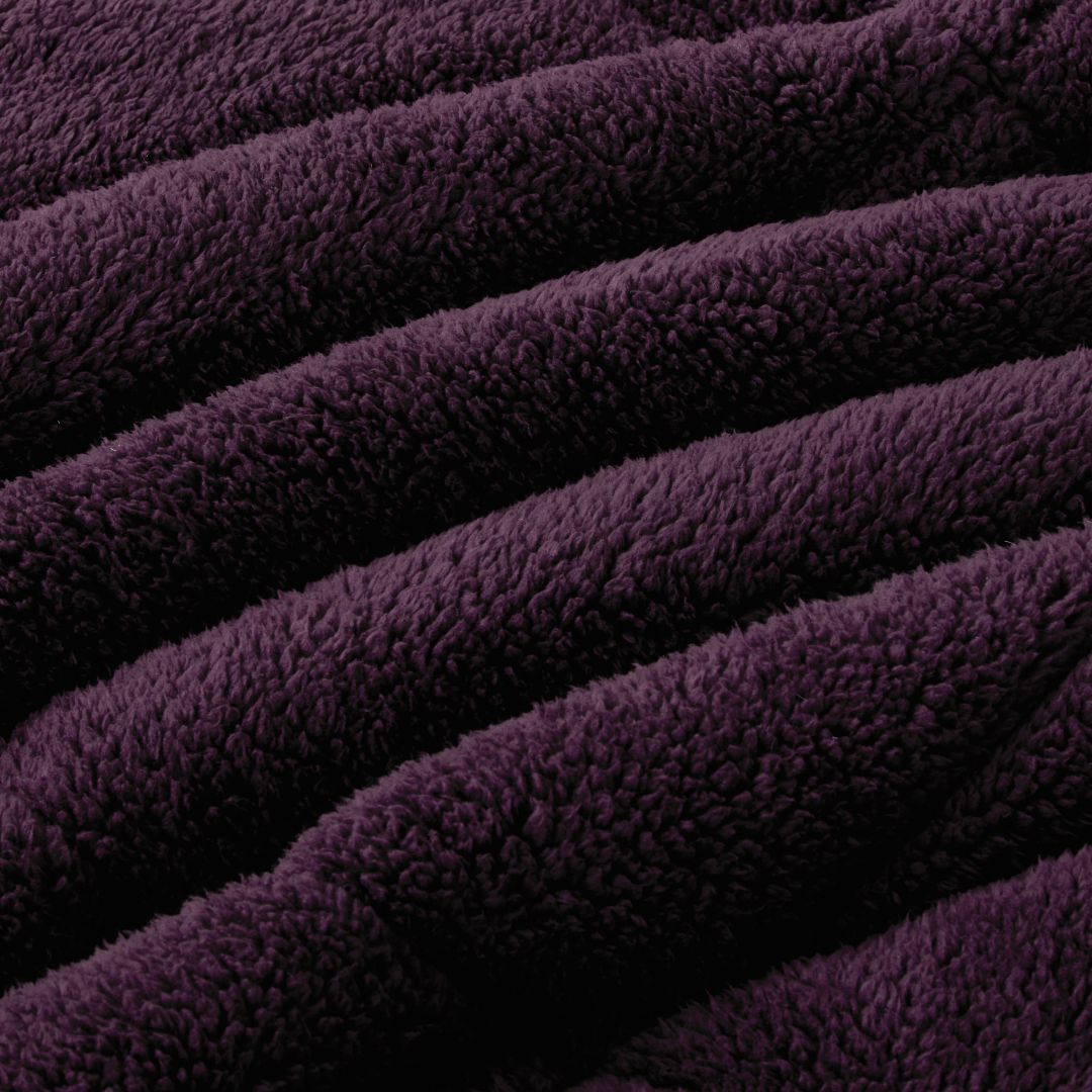 A neatly made bed with the Morgan and Reid Plum Snuggle Fleece Comforter Set and pillows sits on a light rug. Pendant lights hang above, with a side table, vase, and a large potted plant against a textured beige wall.