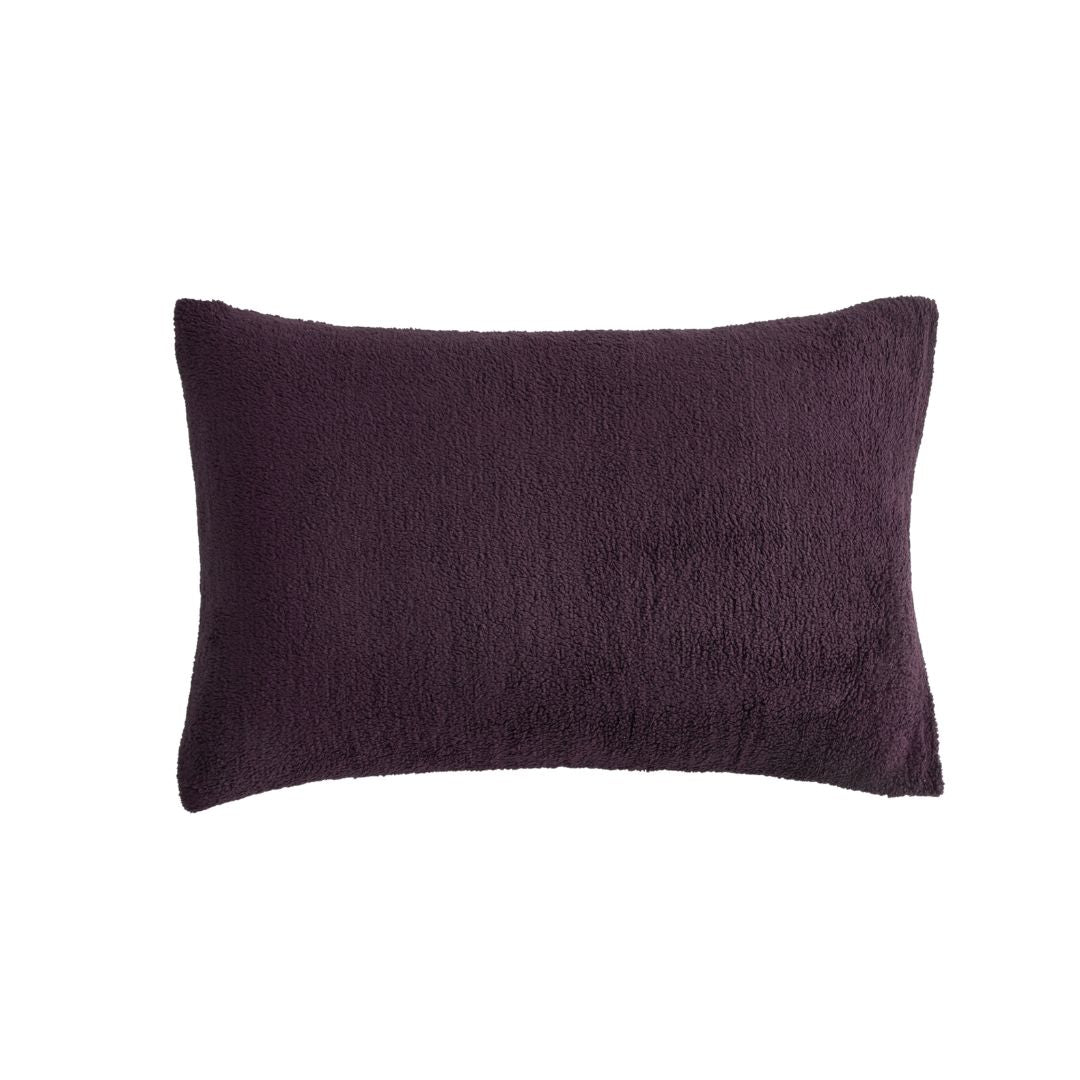 A neatly made bed with the Morgan and Reid Plum Snuggle Fleece Comforter Set and pillows sits on a light rug. Pendant lights hang above, with a side table, vase, and a large potted plant against a textured beige wall.