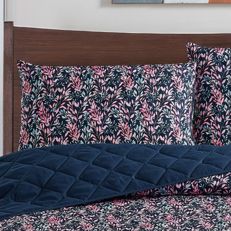 A neatly made bed with a dark floral comforter and matching pillows, the Morgan and Reid Sophia Coverlet Set in lightweight breathable fabric at the foot, a wooden headboard, geometric wall art, and a vase of flowers on the bedside table.