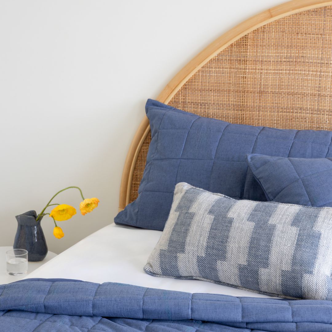 Bed with blue bedding and pillows, wicker headboard, and small vase with yellow flowers on a white surface.