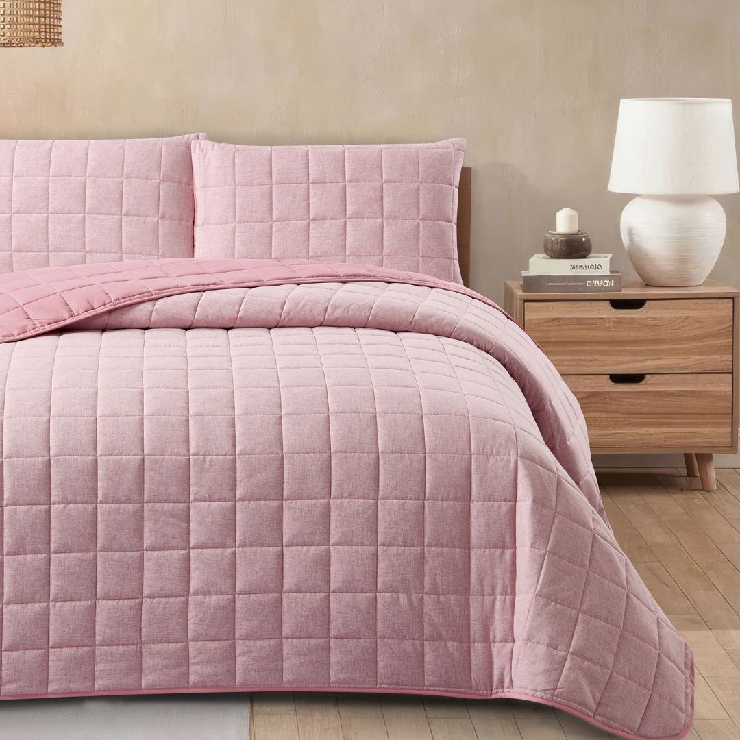 Pink quilted bedding set on a bed with a wooden nightstand and lamp in the background.