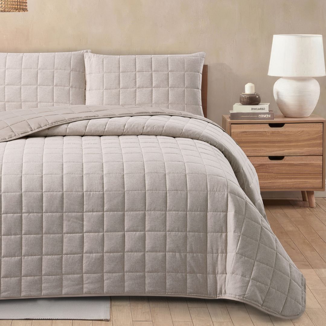 Quilted beige bedspread on a bed with matching pillows, next to a wooden nightstand with a lamp.