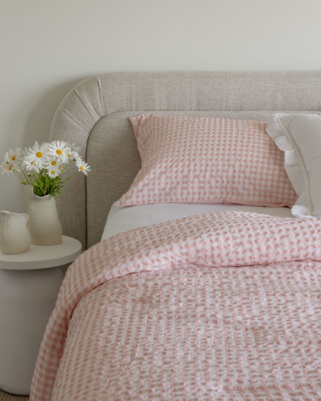 Bed with pink checkered bedding and white flowers on a nightstand.