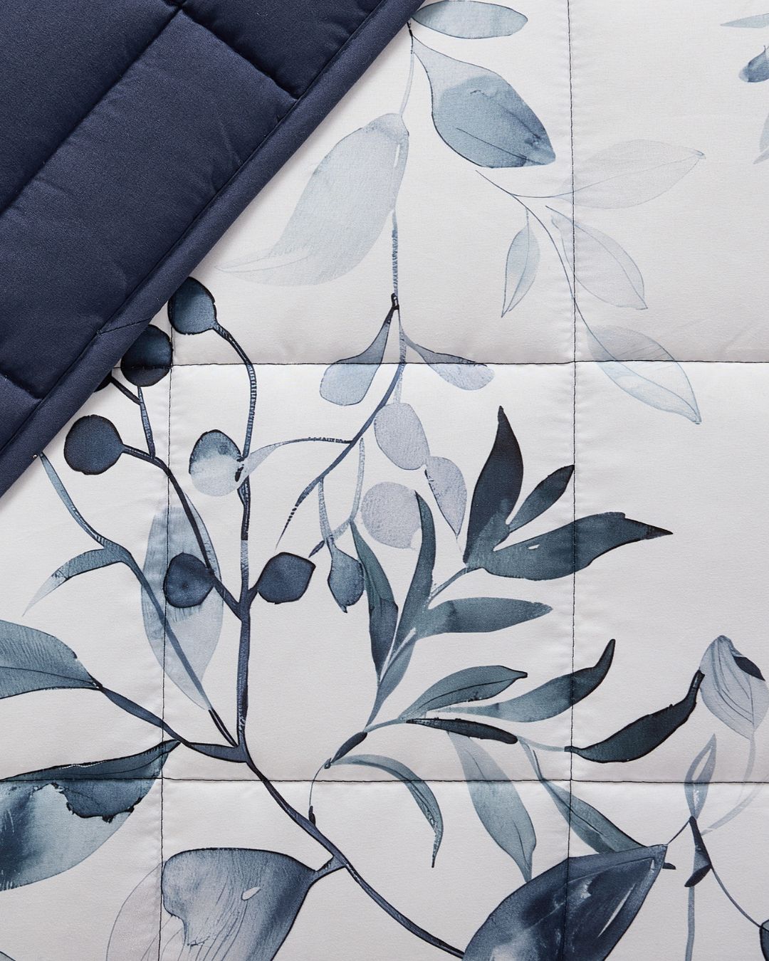 A close-up of a quilted bedding surface with a botanical leaf print in blue-gray and white, with a folded dark navy section at the top left.
