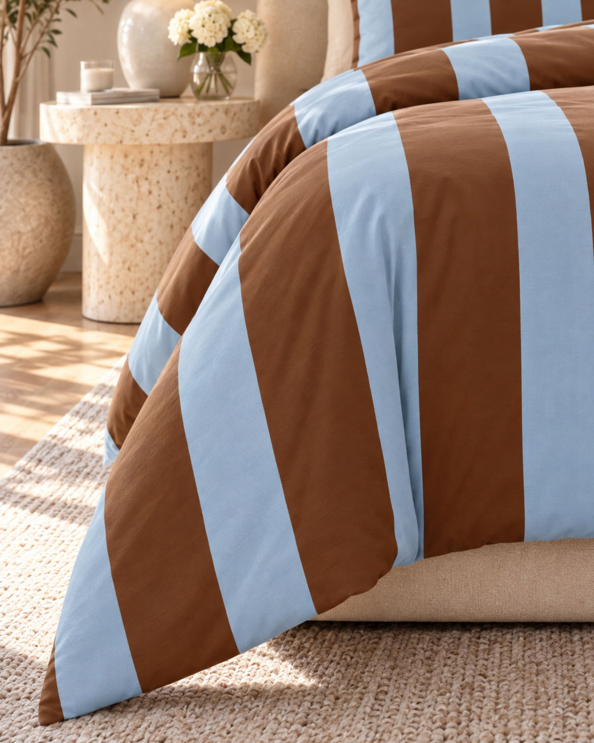 A close-up of a striped bedding set with alternating brown and light blue stripes, styled on a bed in a warm, sunlit bedroom setting.