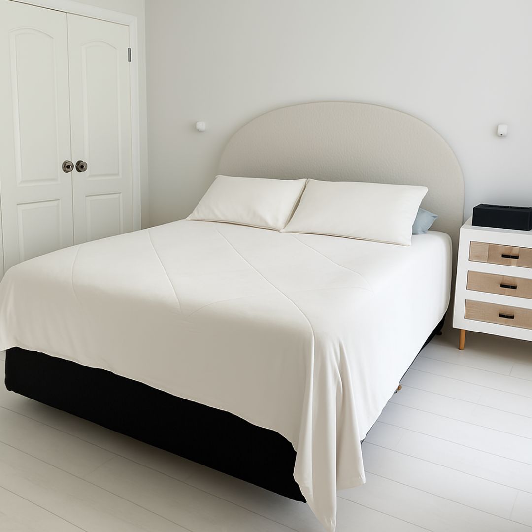 A minimalist bedroom features a neatly made bed with white bedding and two pillows, topped with the Sienna Living Summer Bamboo Cooling 2 in 1 Comforter Quilt. A lamp sits on a small nightstand, and sheer white curtains cover the window.