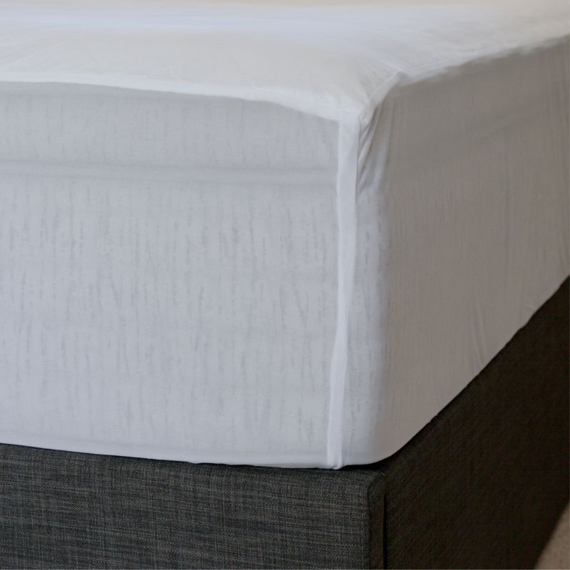 A neatly made bed with white sheets and pillows, a wicker headboard, and the Sienna Living Bamboo Mattress Protector. A small wooden table beside the bed holds a white vase filled with green leafy branches.