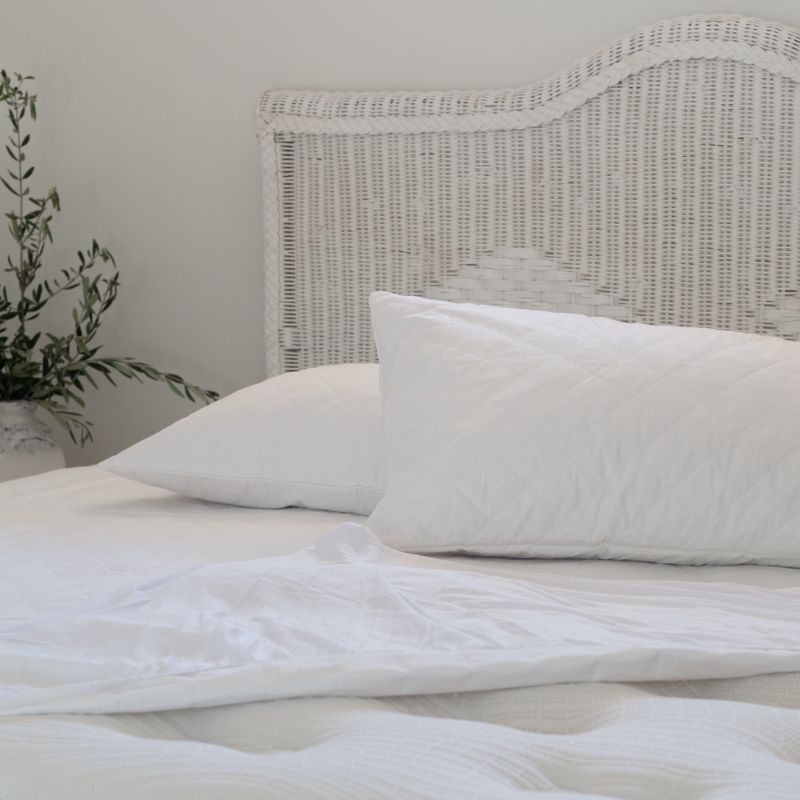 A neatly made bed with white sheets and pillows, a wicker headboard, and the Sienna Living Bamboo Mattress Protector. A small wooden table beside the bed holds a white vase filled with green leafy branches.