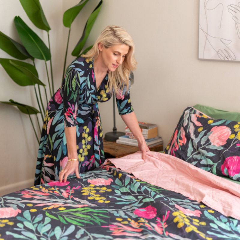 A bed styled with the Sienna Living BSM Claire Coverlet Set—featuring a dark floral pattern with pink flowers and green leaves—plus matching pillows, wooden nightstands, plants, and minimalist art on the white wall above.