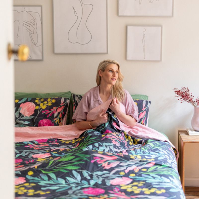A bed styled with the Sienna Living BSM Claire Coverlet Set—featuring a dark floral pattern with pink flowers and green leaves—plus matching pillows, wooden nightstands, plants, and minimalist art on the white wall above.