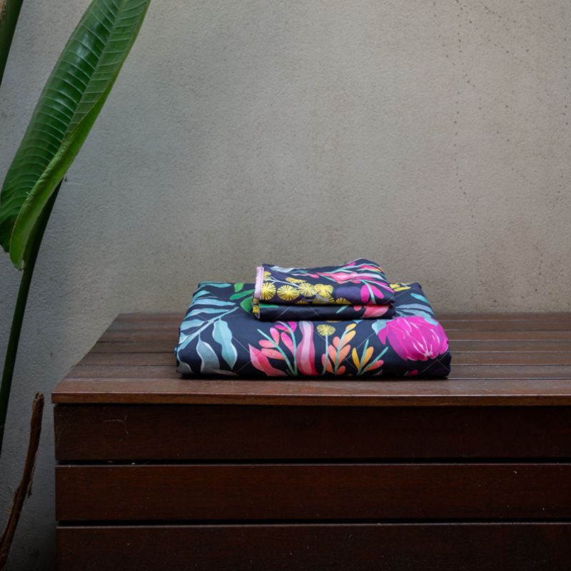 A bed styled with the Sienna Living BSM Claire Coverlet Set—featuring a dark floral pattern with pink flowers and green leaves—plus matching pillows, wooden nightstands, plants, and minimalist art on the white wall above.