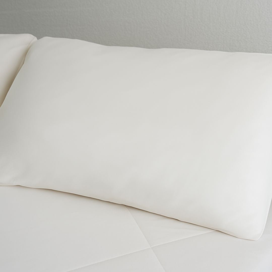 A neatly made bed with white pillows and Sienna Living Cooling 2 in 1 Pillowcase Pair sits beside a white nightstand with a black tissue box and beige bowl, all set against a minimal white wall for a refreshing sleep surface.
