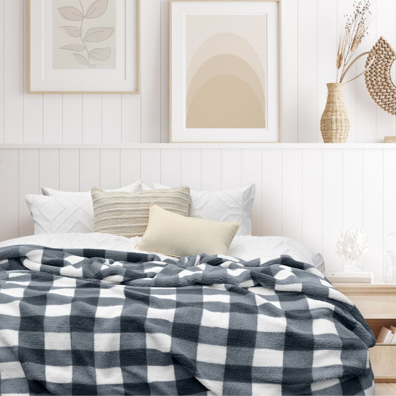 A cozy bed with white pillows and a black-and-white plaid blanket, set in a bright room with neutral decor, framed art, and woven accents on the wall and nightstand.