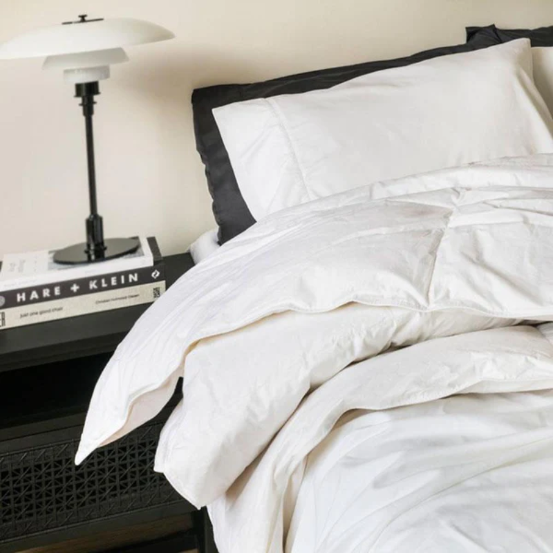 A neatly made bed with white bedding and pillows is beside a black nightstand holding two stacked books and a black and white modern lamp.