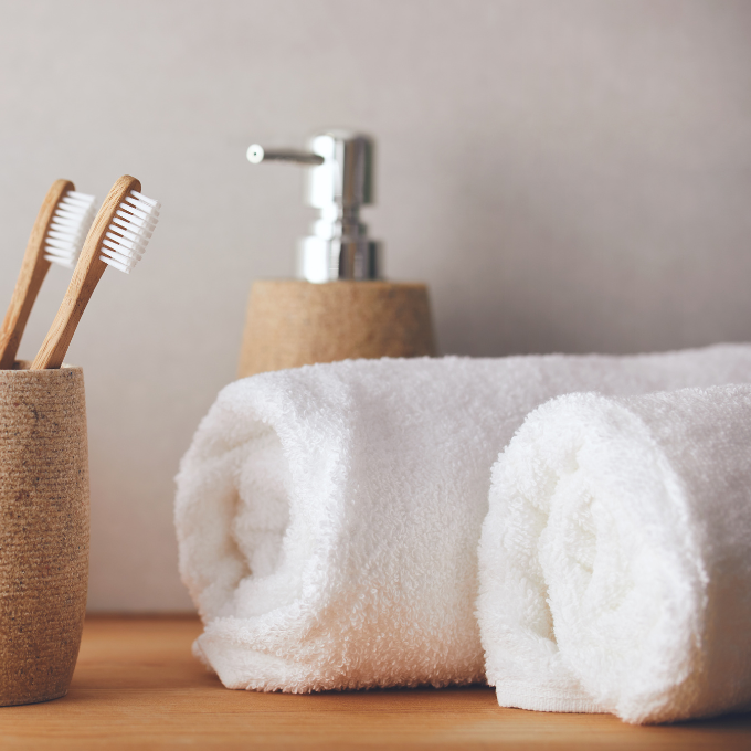 Two rolled white towels, a soap dispenser, and two wooden toothbrushes in a cup are arranged on a wooden surface against a light, neutral background.
