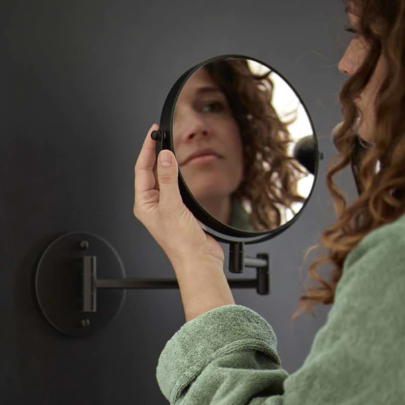 A woman in a green robe looks at her reflection in a round, wall-mounted magnifying mirror against a dark background.