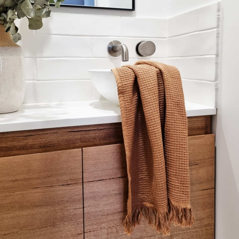 A brown textured hand towel with fringed edges hangs over a white vessel sink on a wooden bathroom vanity, with white tile backsplash and a vase with greenery nearby.
