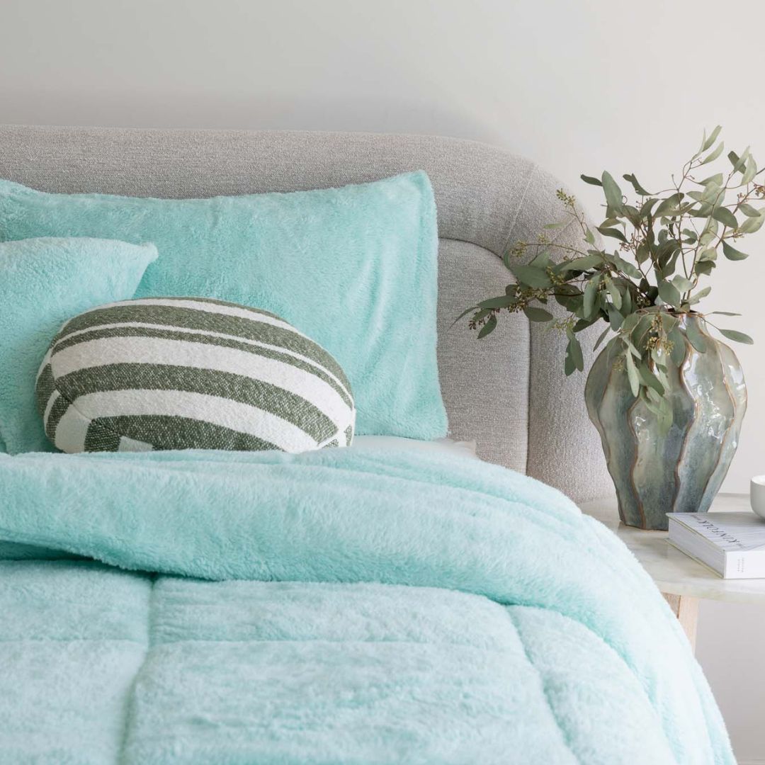 A modern bedroom with a white bed frame, mint green bedding and pillows, a round white side table with glass decor, a potted plant, and neutral walls with a built-in shelf.