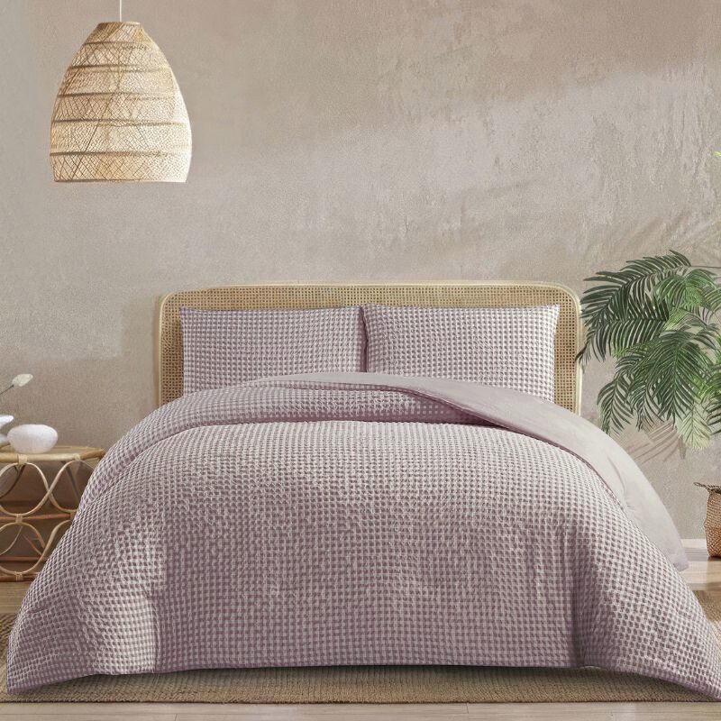 A bed with a light wood headboard is made up with a green and white buffalo check comforter set. The room has white brick walls, a geometric pendant light, and minimalist decor.