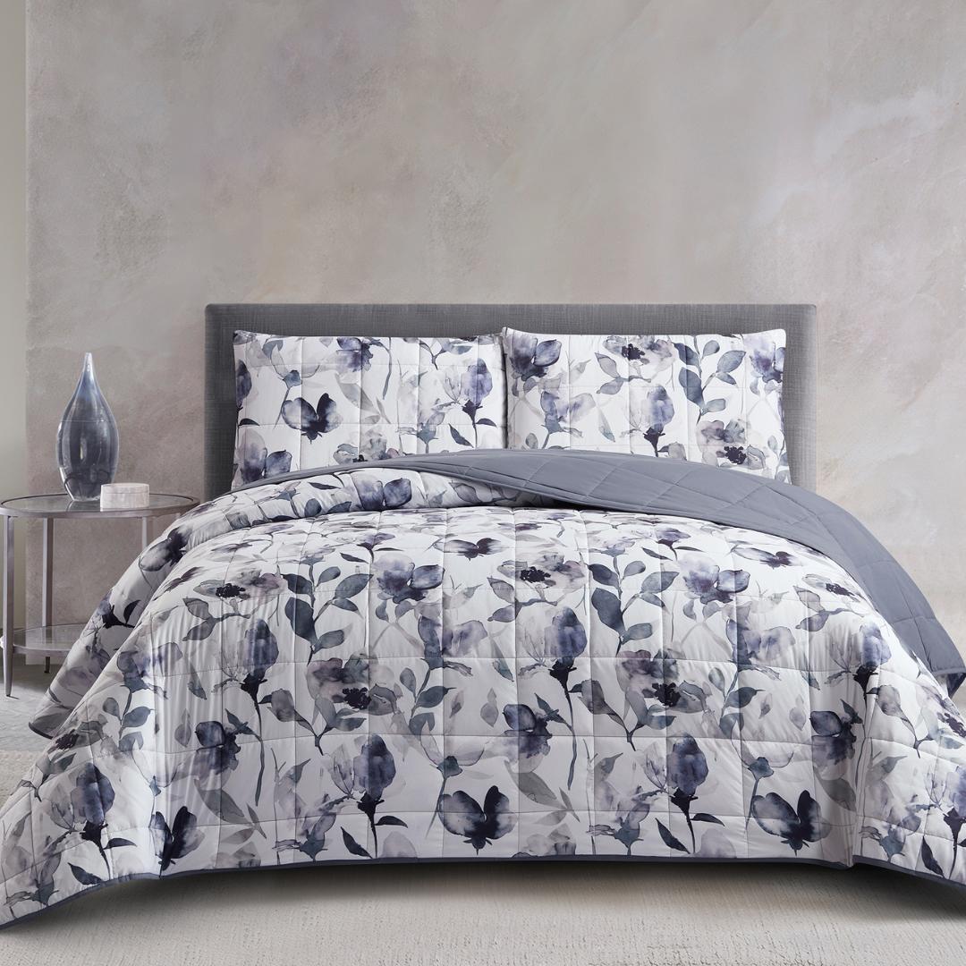 A neatly made bed with gray floral-patterned bedding and matching pillow shams, next to a round nightstand with a glass vase, against a light gray textured wall.