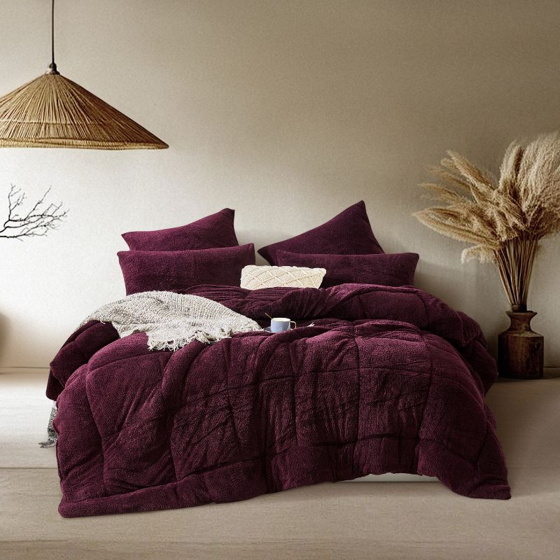 A cozy bed with deep purple bedding, large pillows, a knit throw, and a coffee cup sits in a neutral-toned room with pampas grass and a woven pendant light as decor.