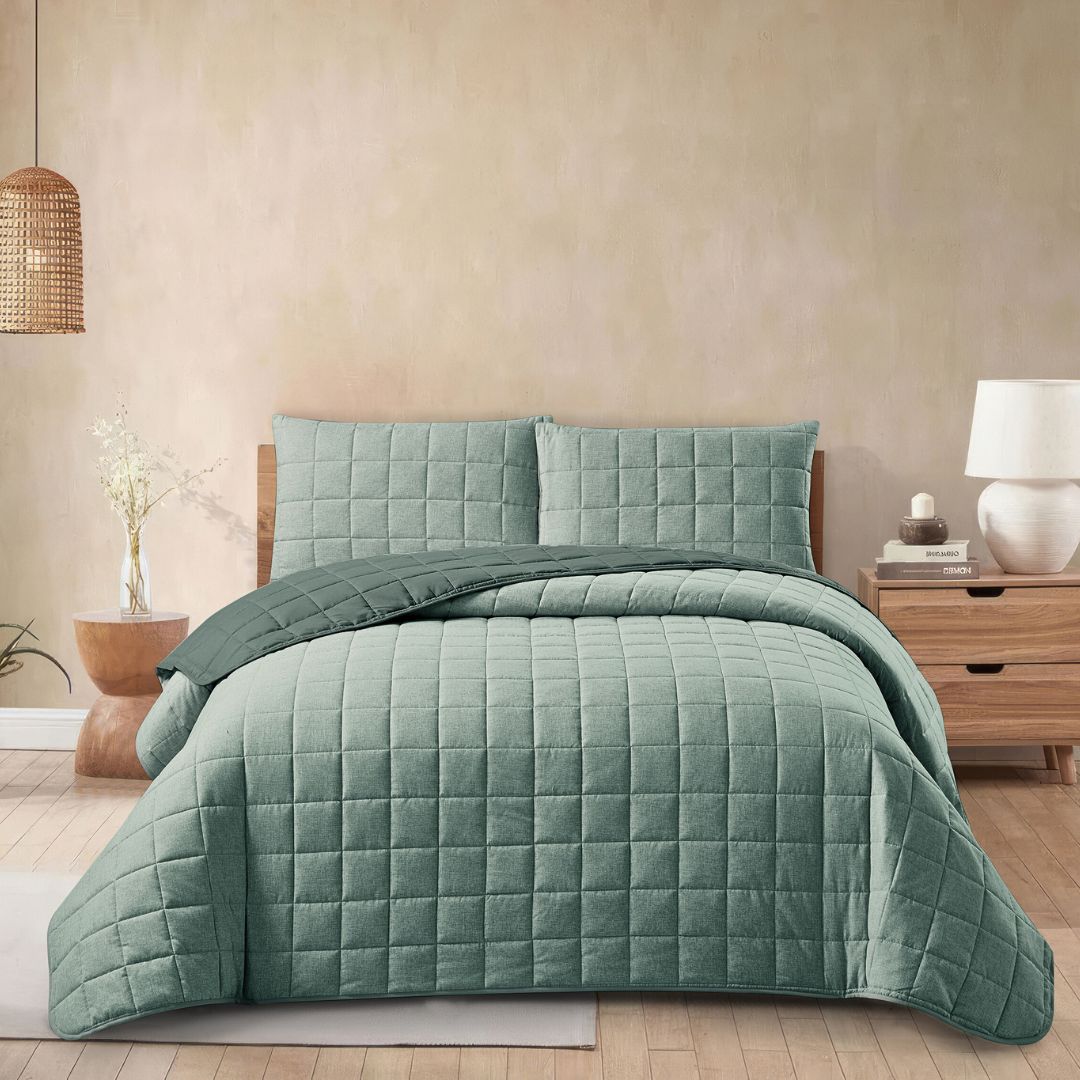 A neatly made bed with a greenish-blue quilted comforter and pillows in a cozy bedroom with a wooden nightstand, lamp, vase, and woven pendant light.