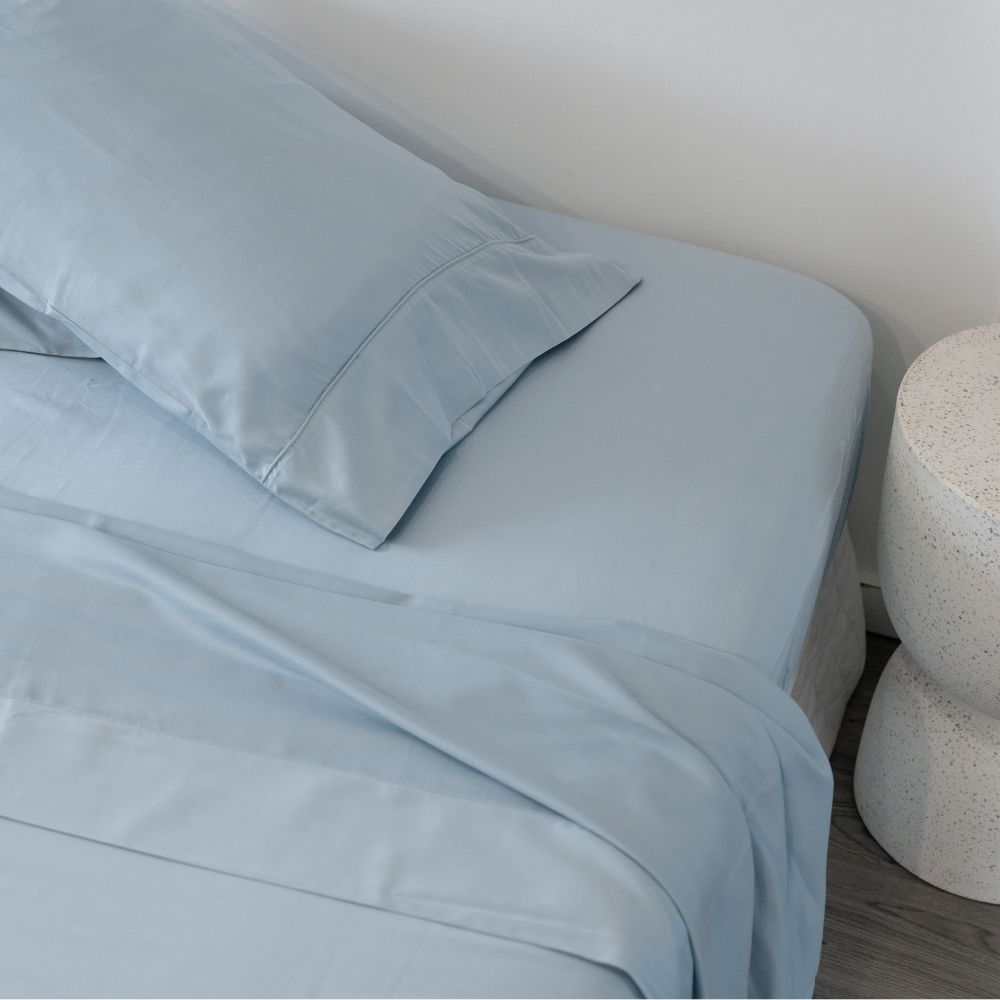 Light blue bed sheets and pillowcase on a neatly made bed next to a small round white bedside table, with a gray wooden floor partially visible.